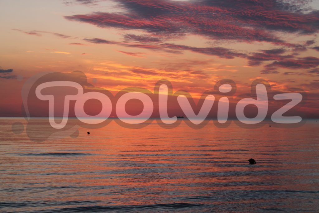 Vista mare con l'alba all'orizzonte e una piccola figura di una barca adibita alla pesca. Riflesso arancione e rosso sulla distesa d'acqua.