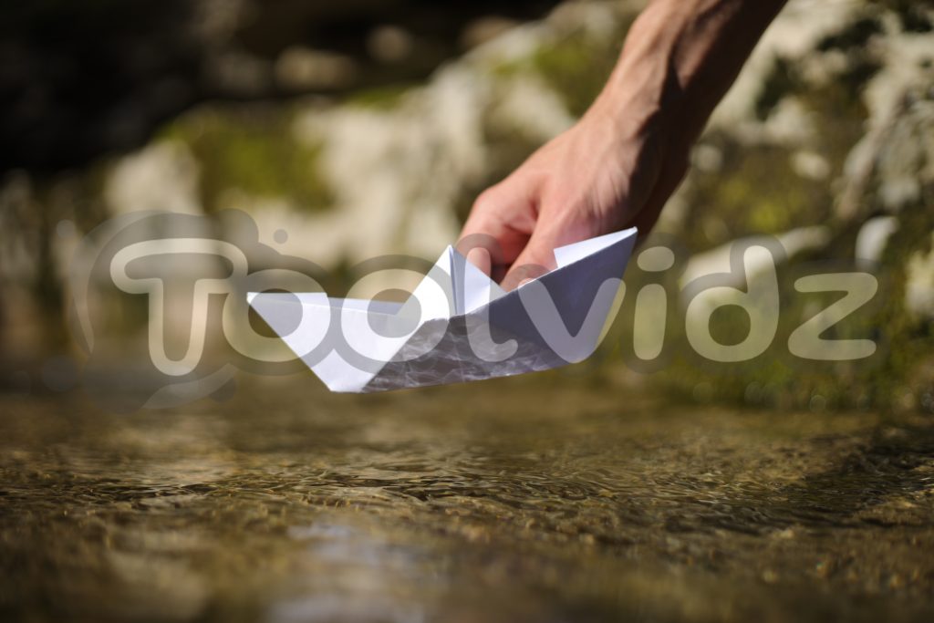 Una mano di un ragazzo giovane sta per poggiare una barca di carta su un telo d'acqua di torrente
