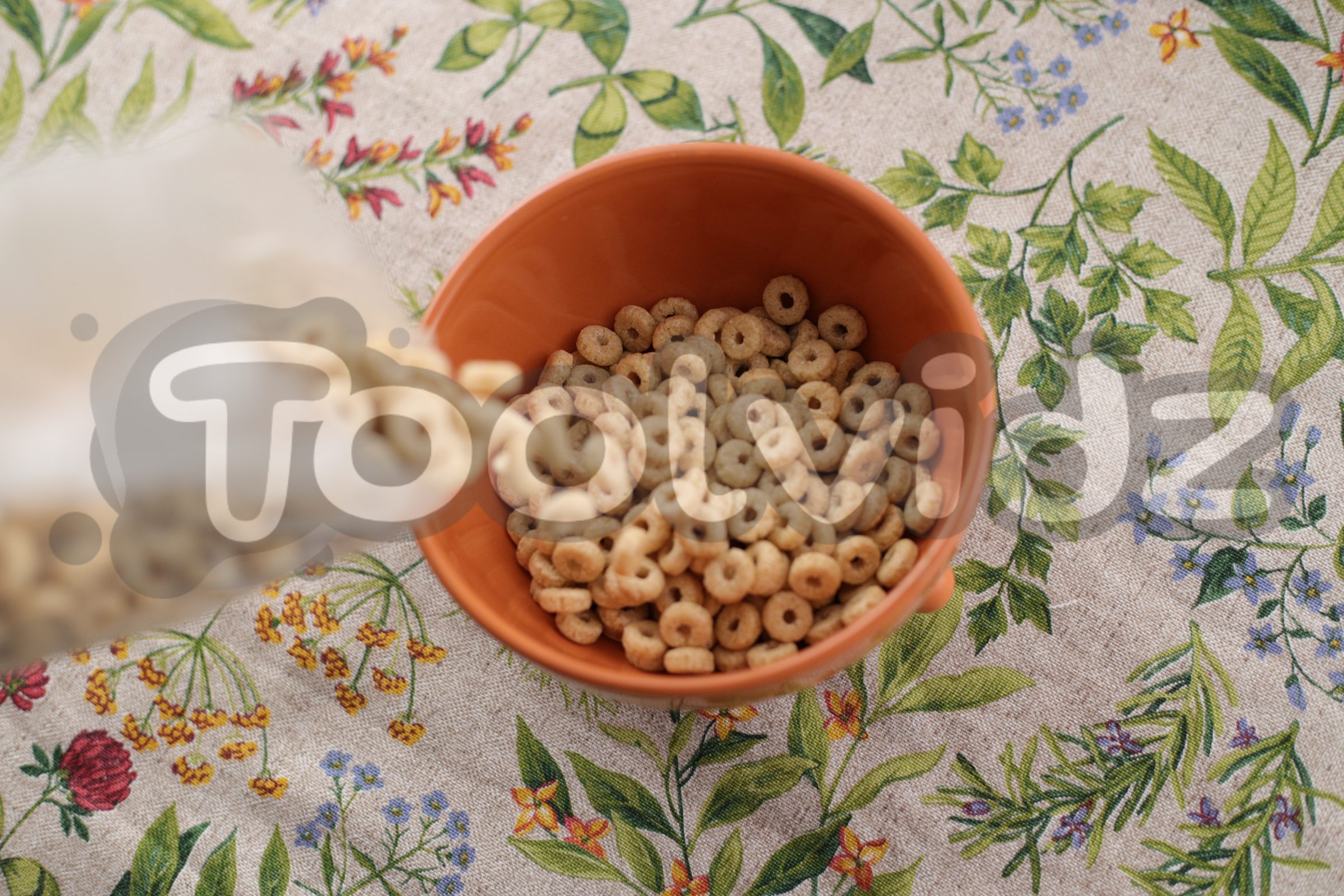Una tazza vista dall'alto, poggiata su una tovaglia floreale, si sta riempiendo ancor di più di cereali